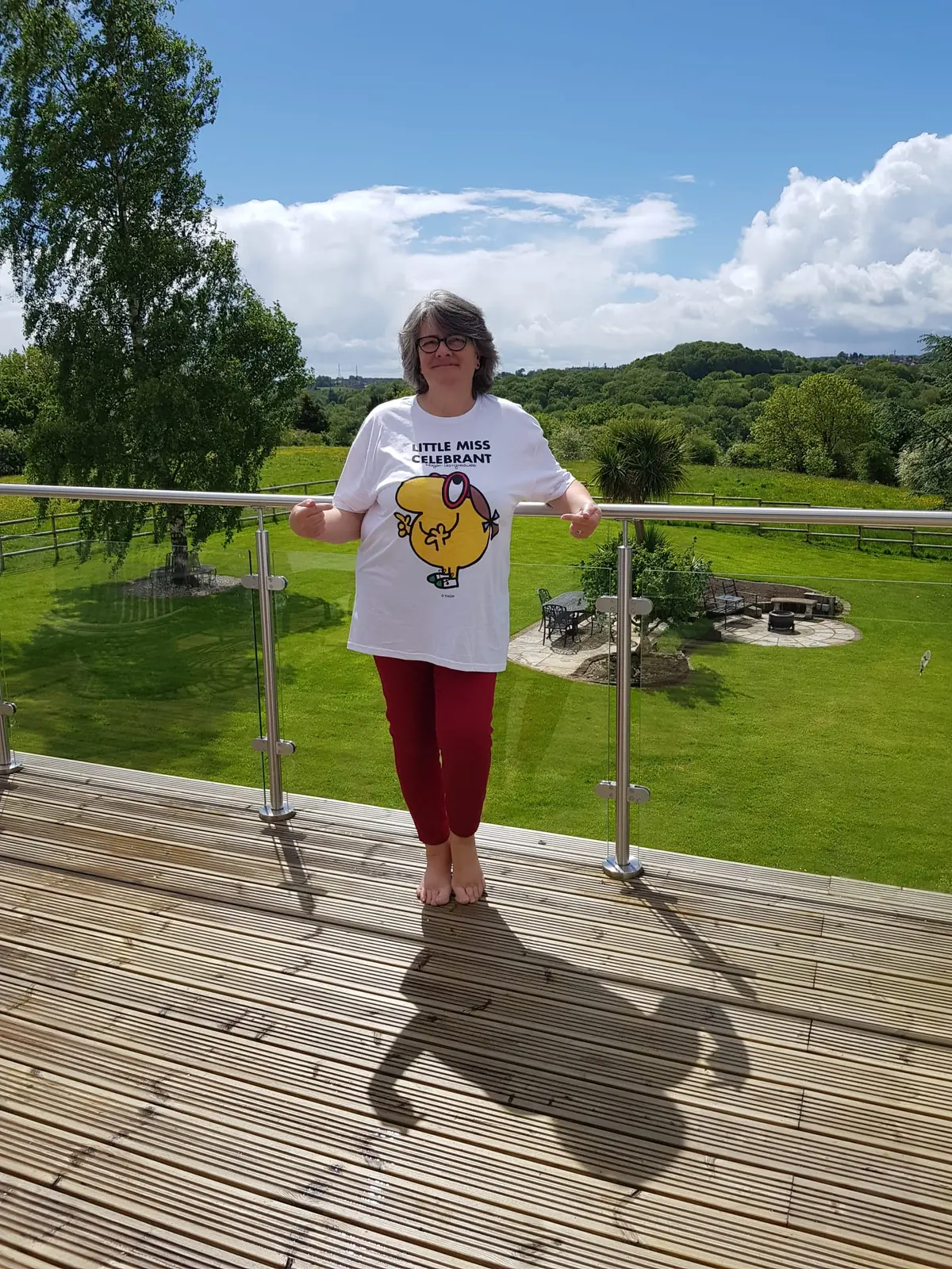 Marian standing barefoot on the sunny decking at The Woodlands, wearing glasses and a white Little Miss Celebrant t-shirt, with the lawn and wooded Shropshire hills behind her.