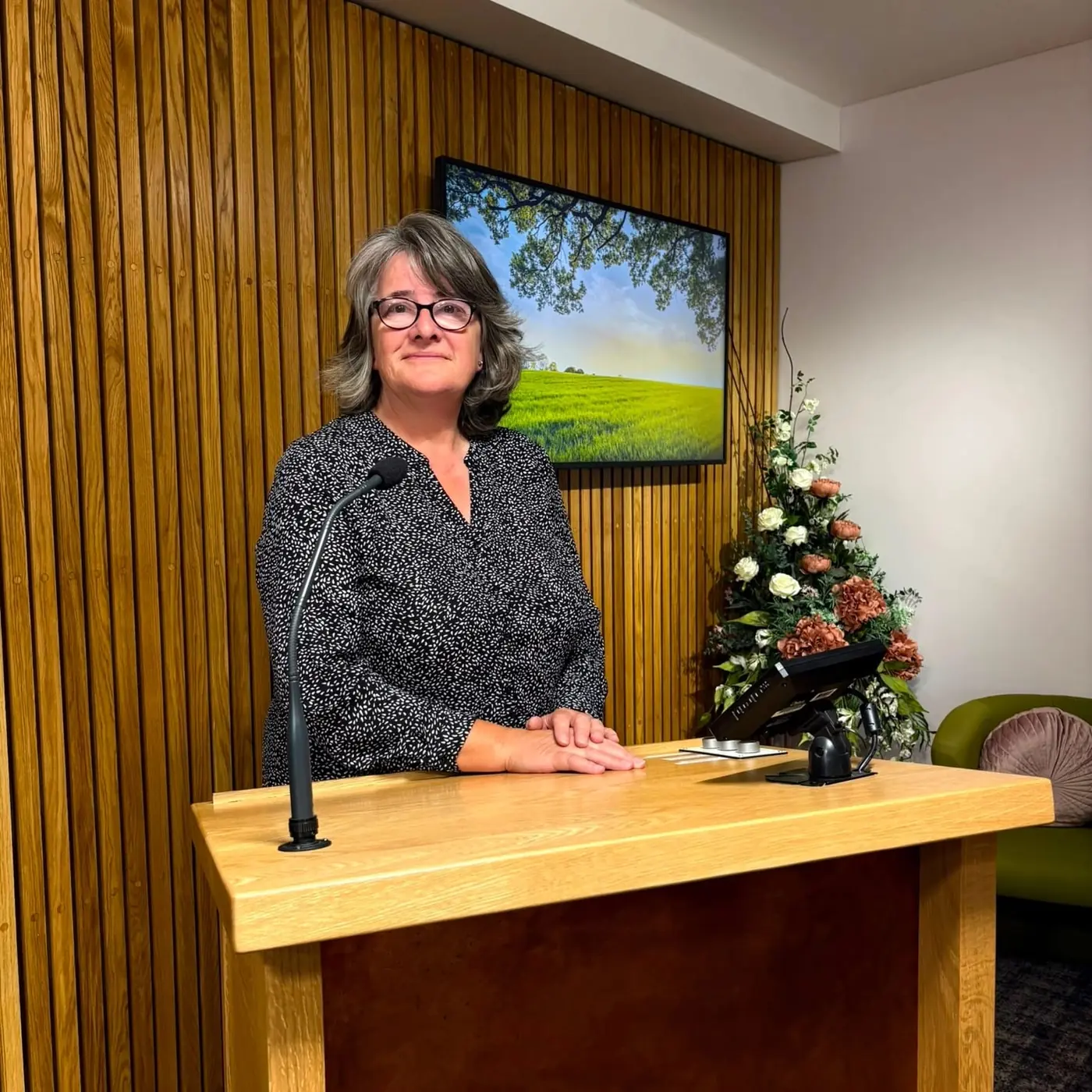 Marian standing calmly at a light oak lectern in a wood-panelled crematorium, her hands resting together, with flowers arranged beside her.